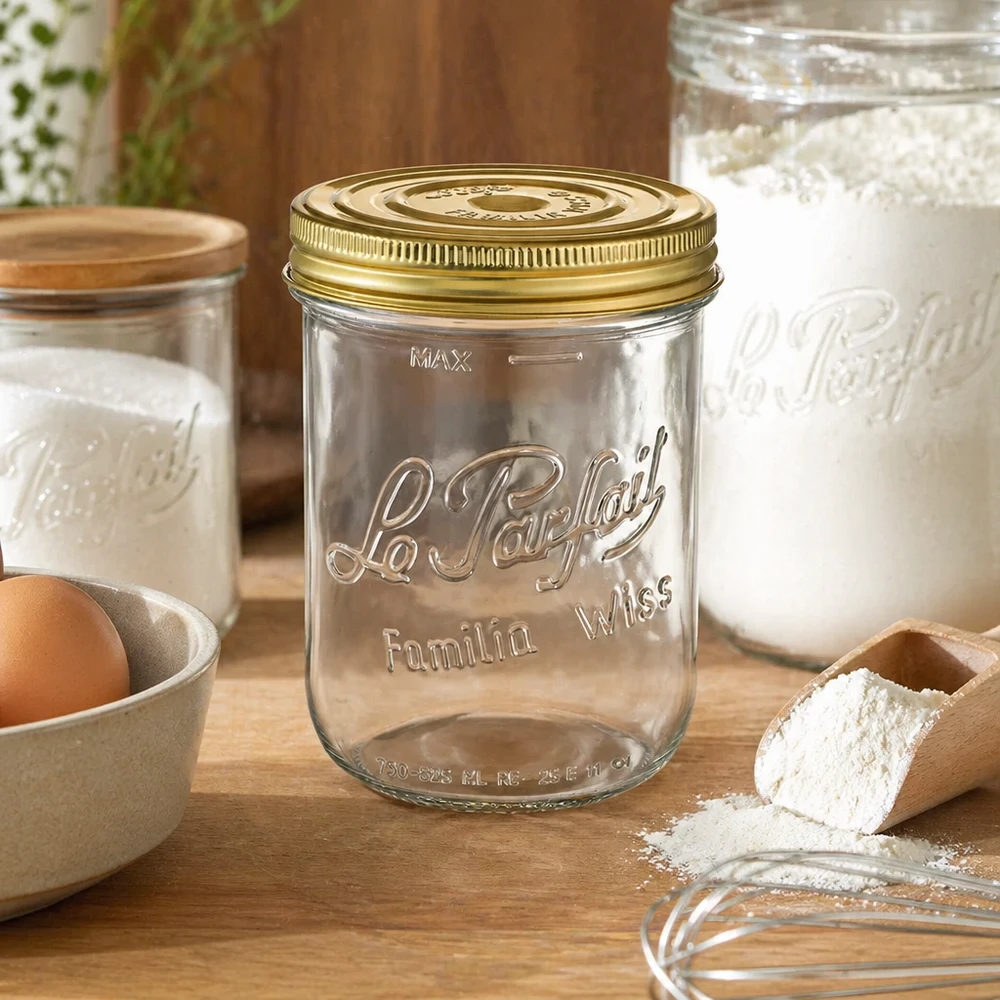 Le Parfait® Familia Wiss 1 kg Ø100 mm Bocal Pot de 1020 ml Familia Wiss avec couvercle doré sur une table en bois à côté d'ingrédients de pâtisserie et d'autres bocaux.