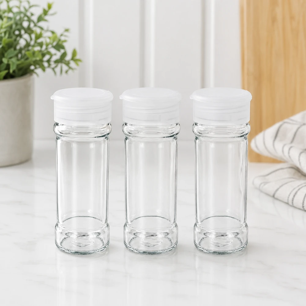 Trois bocaux à épices en verre transparent vides de 100 ml avec des couvercles saupoudreurs blancs, posés sur une surface en marbre